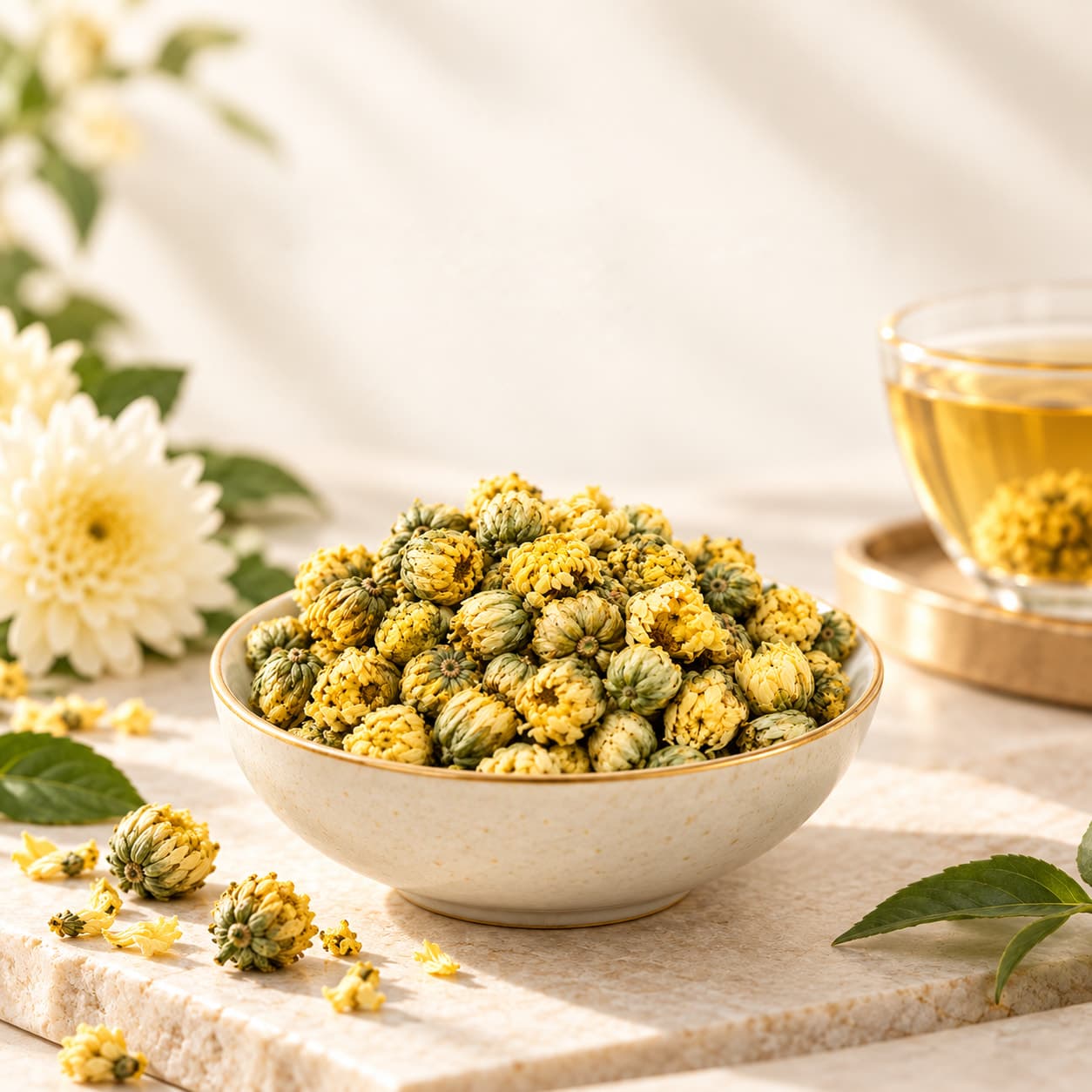 Dried chrysanthemum buds heaped in a stoneware bowl on white stone, white chrysanthemums and a cup of infusion behind.